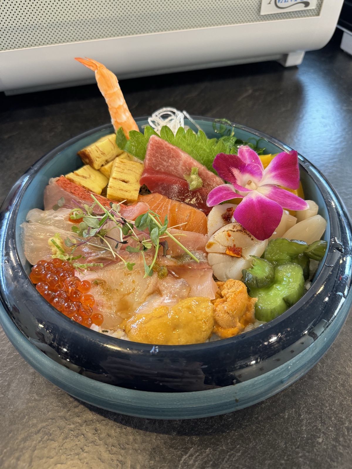 Chirashi don in a blue ceramic bowl: assorted sashimi, shrimp, uni, ikura, and tamago over rice, garnished with an orchid.