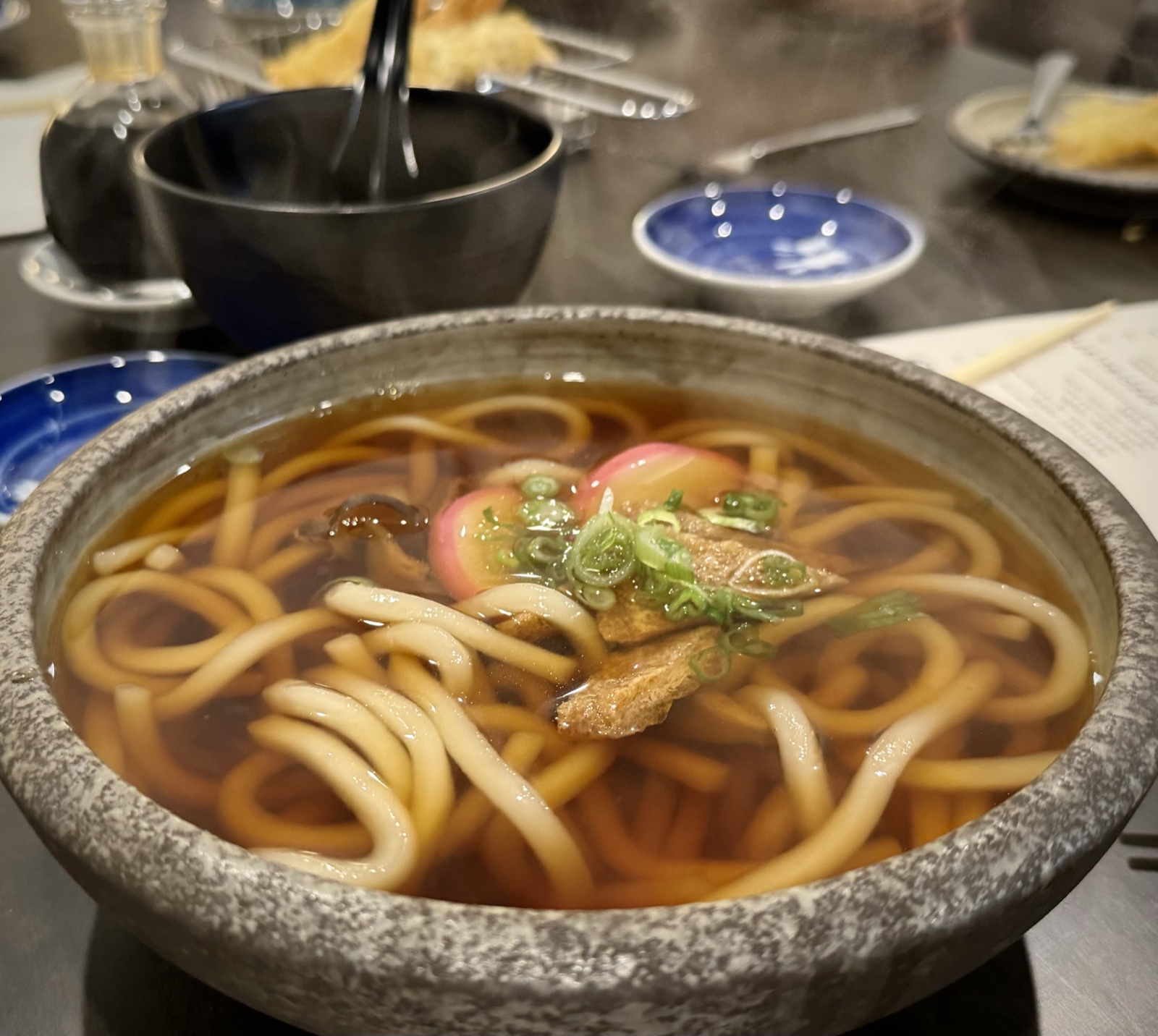 Steaming udon in dark broth in a stone bowl, topped with kamaboko, scallions, and tempura on the side.