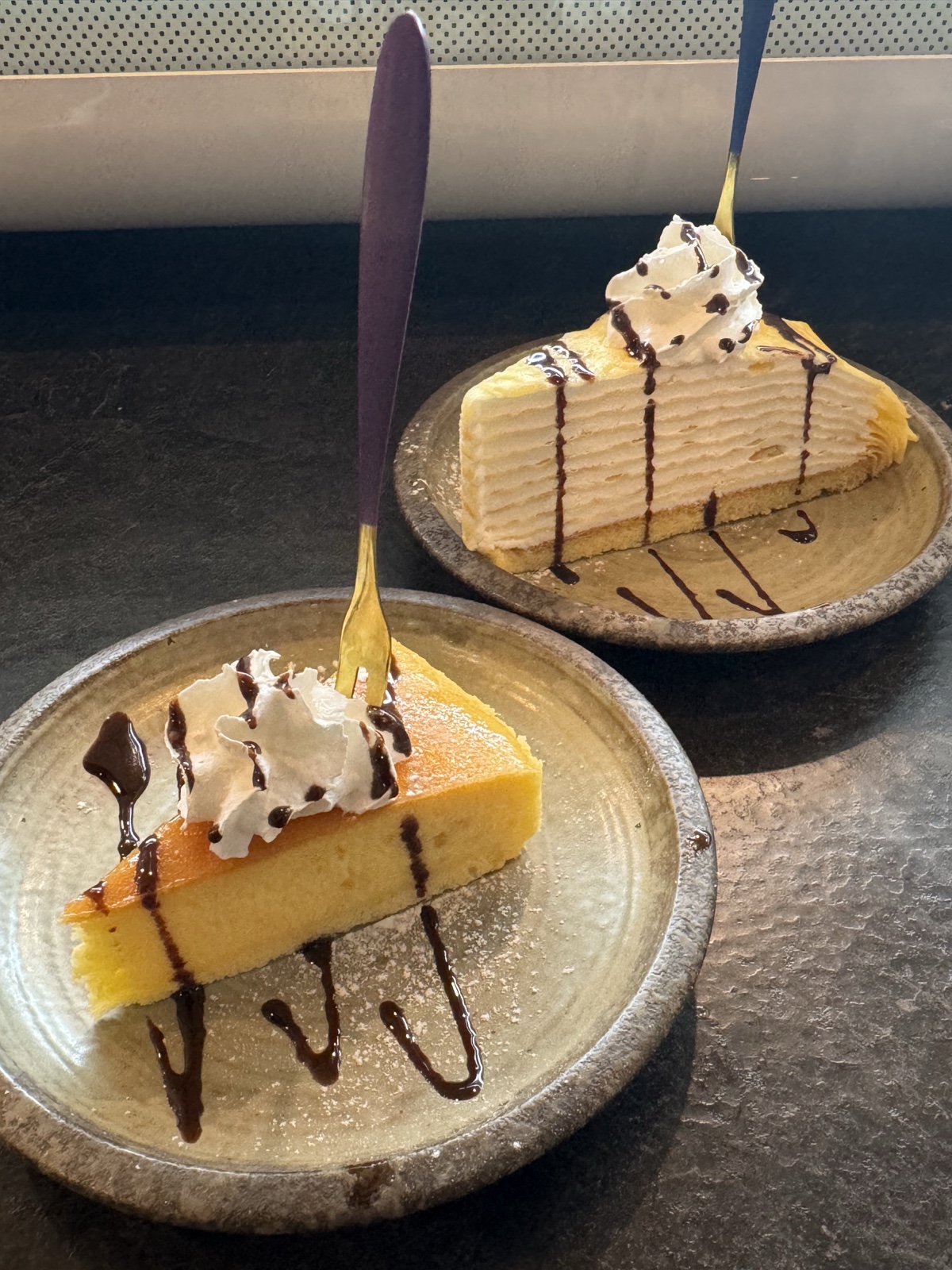 Two dessert slices on stoneware plates with whipped cream and chocolate drizzle, yuzu cheesecake in front.